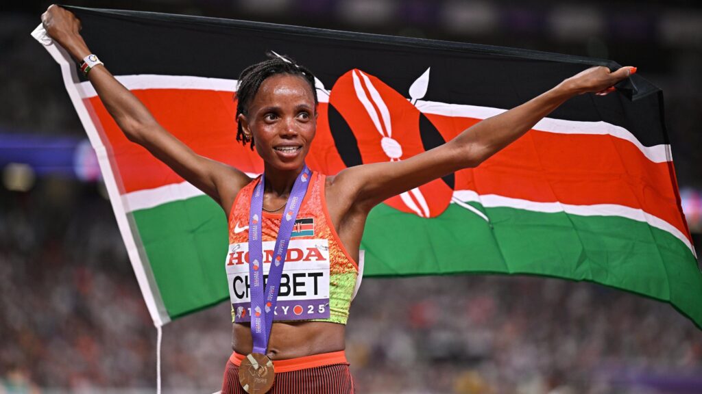 Beatrice Chebet wears a gold medal on a purple ribbon over an orange and green Kenya running vest as she holds a Kenyan flag aloft behind her. She has a smile on her face as she celebrates winning the 10,000m at the World Athletics Championships in Tokyo