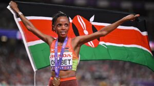 Beatrice Chebet wears a gold medal on a purple ribbon over an orange and green Kenya running vest as she holds a Kenyan flag aloft behind her. She has a smile on her face as she celebrates winning the 10,000m at the World Athletics Championships in Tokyo