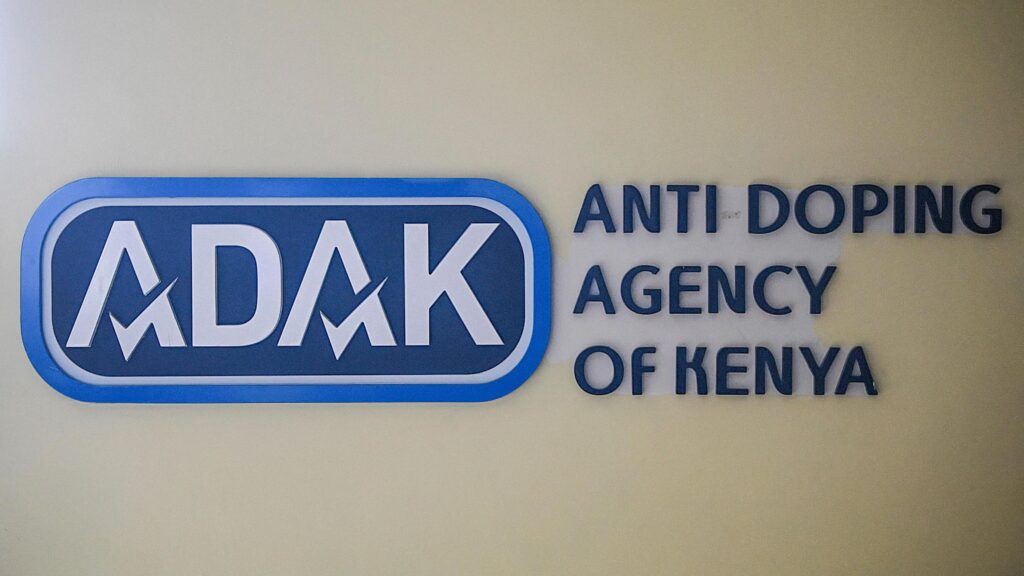 The blue and white logo of the Anti-Doping Agency of Kenya, ADAK, is displayed on a wall with the full text of the organisation's name to the right of the logo