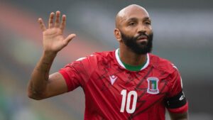 Emilio Nsue, wearing a red Equatorial Guinea shirt with a number 10 on the chest, holds his right hand aloft during a football game. The player has a bushy black beard and a neutral expression on his face