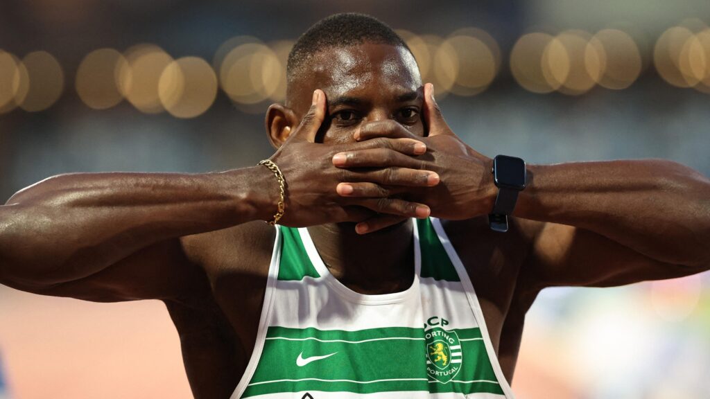 Emmanuel Eseme, seen from chest up wearing a vest with green and white horizontal stripes and a Sporting Clube de Portugal crest on it, covers his mouth with interlocking fingers to mimic the goal celebration of Arsenal striker Viktor Gyokeres. A row of bright lights and an athletics track are seen blurred in the background
