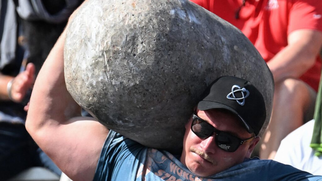 Rayno Nel of South Africa seen from chest up, wearing a blue t-shirt, black cap and black sunglasses, performs the stone to shoulder press at 2025's World Strongest Man in Sacramento. He has a look of concentration and effort on his face as he flexes his huge bicep and raises his right arm to balance an enormous grey stone in the crook of his neck and against his head. Partially seen in the background are two blurred people, one of whom is clapping