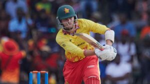 Brian Bennett, wearing a green cricket helmet with a Zimbabwe crest on the front, a yellow Zimbabwe shirt, red trousers and red pads, is seen holding a bat while playing a cricket shot on the front foot. Blue stumps with orange bails are seen behind him, with a crowd out of focus in the distance