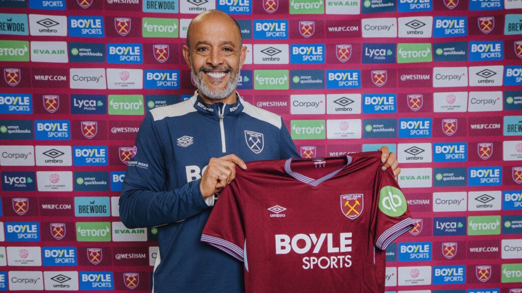 Nuno Espirito Santo, seen from the waist up, smiles during a promo shot for his appointment as the new West Ham United manager. He is wearing a blue West Ham tracksuit top with grey trim, is holding up a claret West Ham home shirt, and is standing in front of a sponsors board with many small rectangular logos on it, including the West Ham club crest
