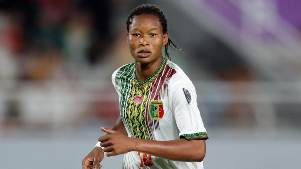 Agueissa Diarra, wearing a white Mali shirt with a colourful green, yellow and red pattern on the front, looks towards the camera as she is seen from waist up running during a football game