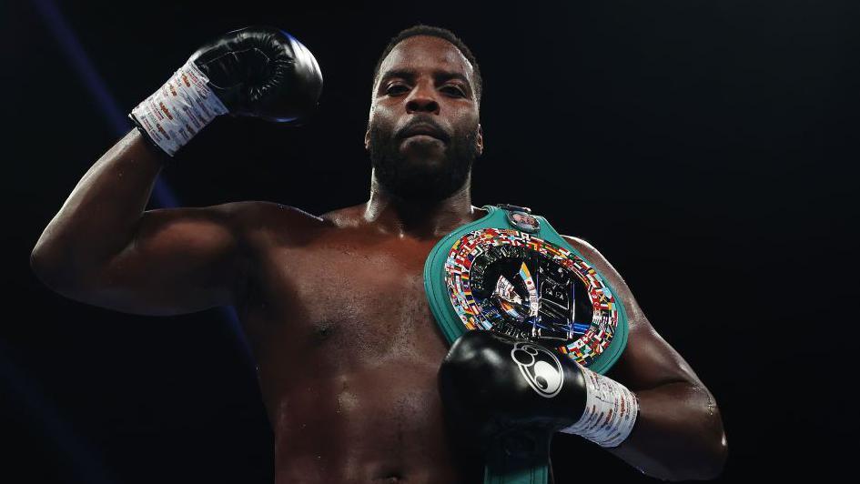 Boxer Lawrence Okolie stands in the ring (which is out of shot) after a fight, posing in a victory stance. Visibly from stomach up, he is bare-chested and muscular. His right arm is bent with his black-gloved fist raised proudly near his head, while his left gloved hand rests on a large championship belt slung over his left shoulder. The belt has a wide teal strap and a shiny, circular centre plate decorated with many small national flags around its edge, catching the arena lights. The background is dark and out of focus, making the boxer and the belt stand out as the clear focal points of the image