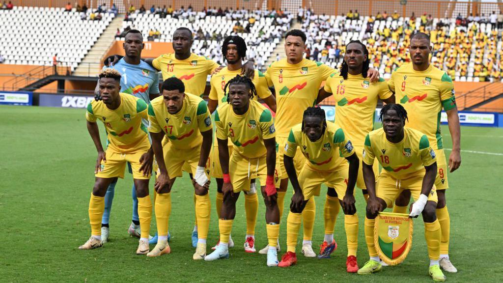 Ten Benin players, all wearing yellow shirts yellow shorts and yellow socks, and a goalkeeper wearing a sky blue shirt, pose for a team photo ahead of a football match