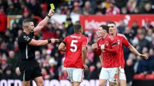 Referee Rob Jones books Elliot Anderson, with the official raising his yellow card above head height and pointing towards the animated Nottingham Forest player, in red and white, who has both hands out in front of his chest