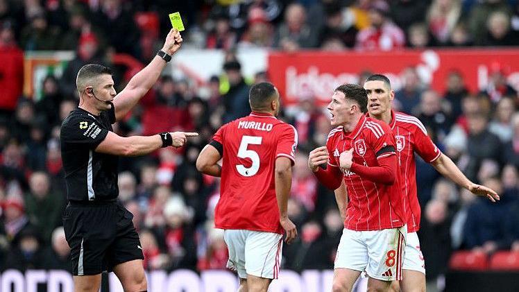 Referee Rob Jones books Elliot Anderson, with the official raising his yellow card above head height and pointing towards the animated Nottingham Forest player, in red and white, who has both hands out in front of his chest
