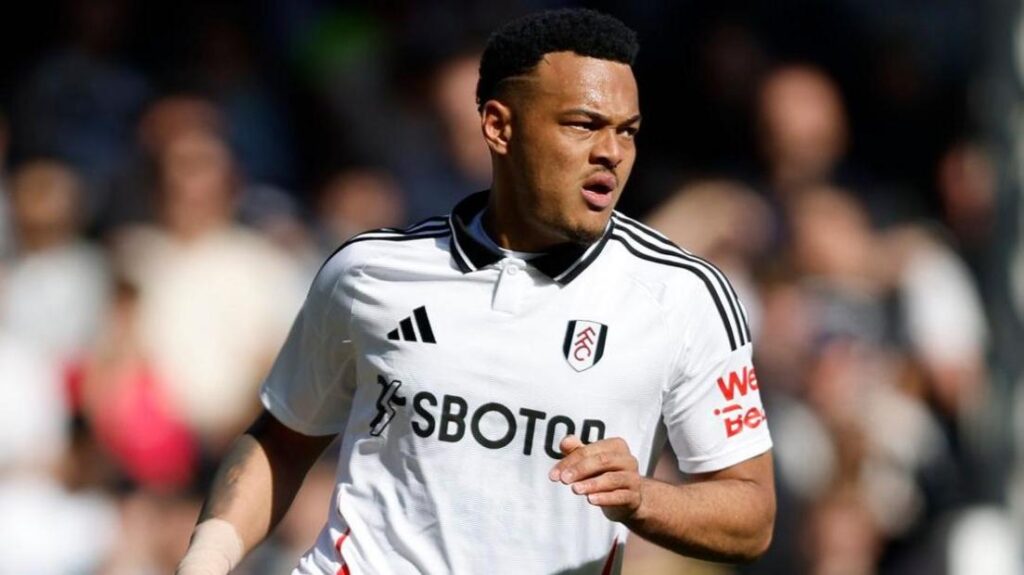 Rodrigo Muniz playing for Fulham. It looks like he is running and leaning to his left. His mouth is open. He is wearing a white Fulham home kit with the black Adidas logo on the left with the three stripes across the shoulders. The fans in the stand are out of focus behind him.