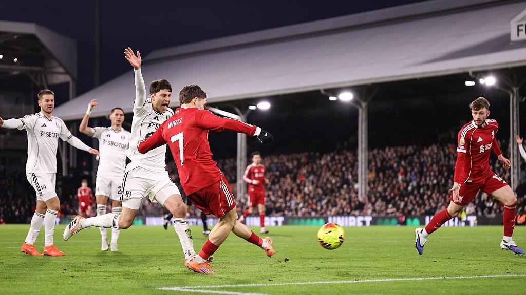 Liverpool's Florian Wirtz scores against Fulham. He is in the penalty area and has struck a yellow ball with his left foot. Liverpool's kit is red and he is wearing orange boots. A Fulham player, wearing all white, is next to him and is raising his right arm to call for offside. Several other players can be seen in the background along with thousands of fans in one of the stands.